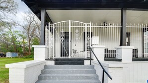 Eine überdachte Veranda mit weißer Brüstung, eine Treppe, die zum Eingang führt, und ein kleines Nebengebäude im Hintergrund.
