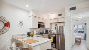 A modern kitchen with white cabinetry, a granite countertop, and stainless steel appliances including a refrigerator, stove, and microwave. There is a small island with a stool, and a dining area visible in the background. - Beach Quarters #2 (Gulfport)