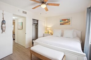 A bedroom with a bed, a bedside table, a lamp, a ceiling fan, a wardrobe, and a painting on the wall. - Beach Quarters #2 (Gulfport)