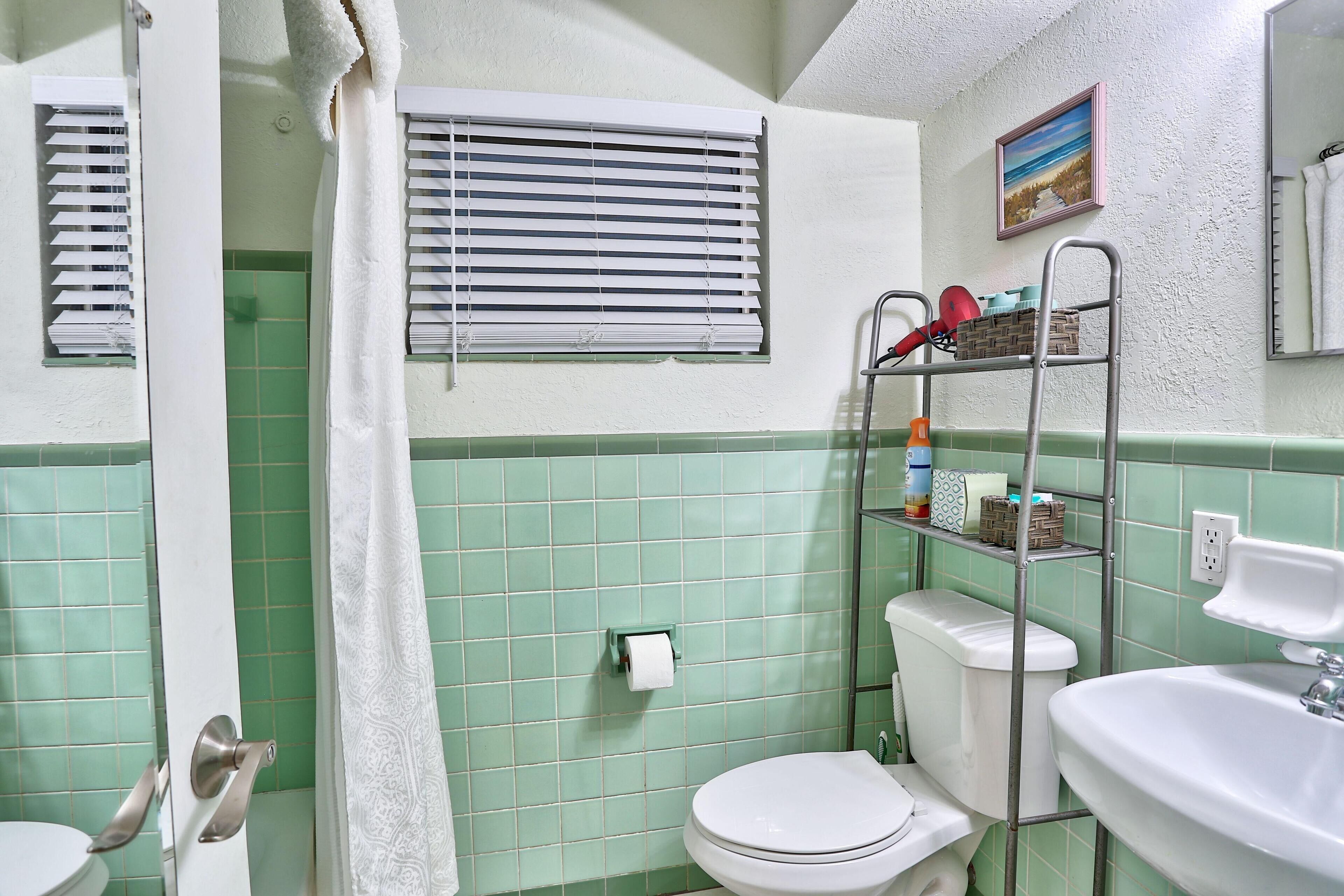Un baño con inodoro, lavabo y zona de ducha. Hay una toalla colgada en la puerta, un perchero y un estante con artículos de aseo.