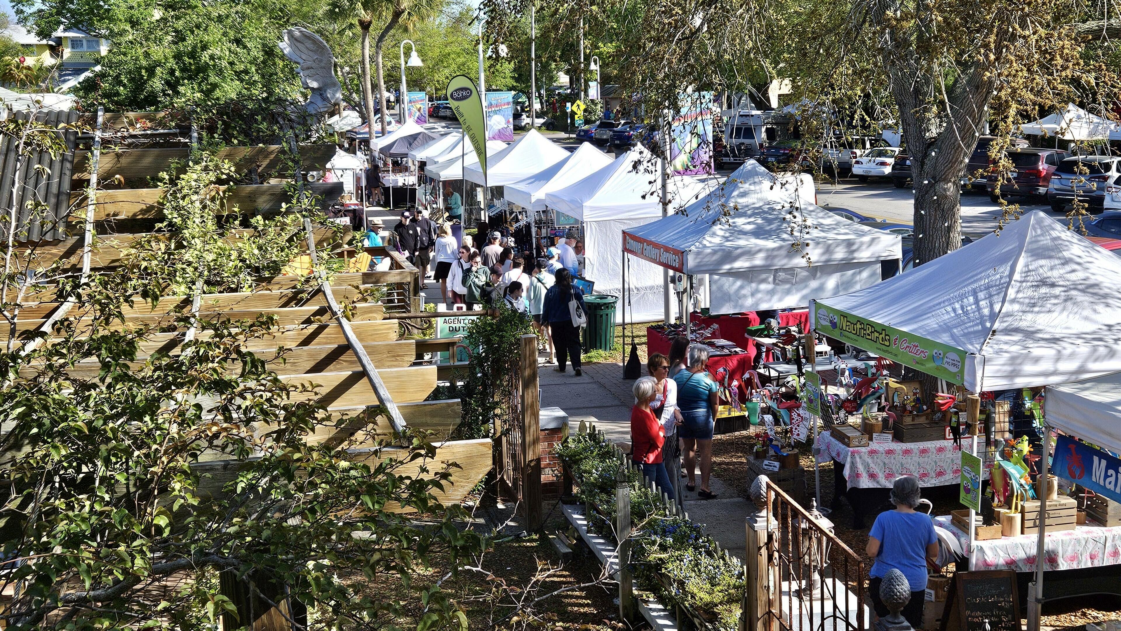 An outdoor market with various stalls and tents, surrounded by trees and a wooden fence.
