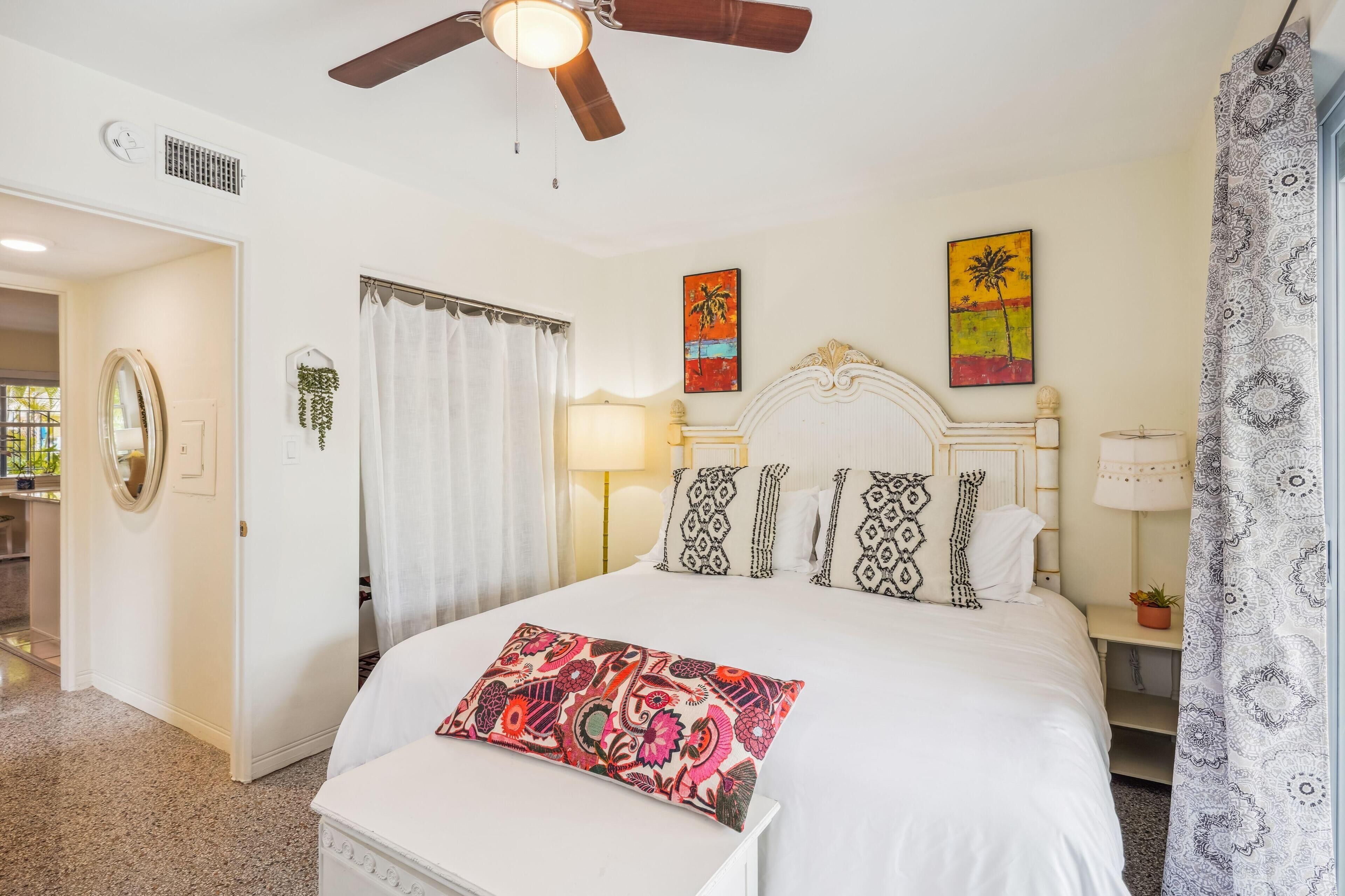 A hotel room with a bed, a ceiling fan, a window with curtains, a bedside table with a lamp, and artwork on the walls.