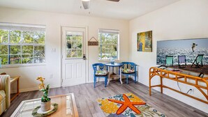 A room with a ceiling fan, two windows with blinds, a door, a small table with chairs, a glass table with chairs, a starfish rug, and a painting of beach chairs. - Beach Quarters #8 (Gulfport)
