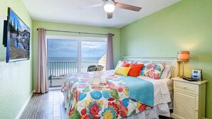 A bedroom with a large bed, a nightstand, a ceiling fan, and a view of the ocean through a sliding door.