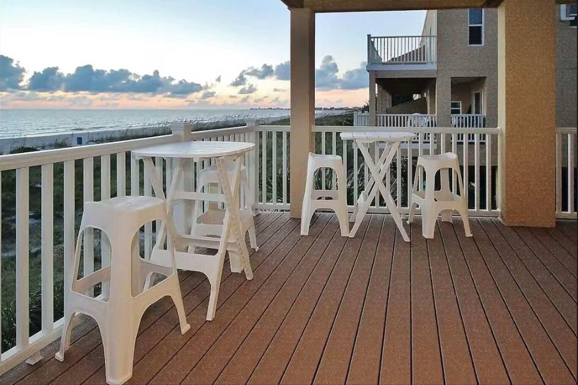 Eine Holzterrasse mit weißen Plastikstühlen und einem Tisch, mit Blick auf ein Gewässer und ein Gebäude mit einem Balkon.