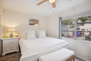 Quarto com cama, mesa de cabeceira, luminária e janela com vista para a rua.