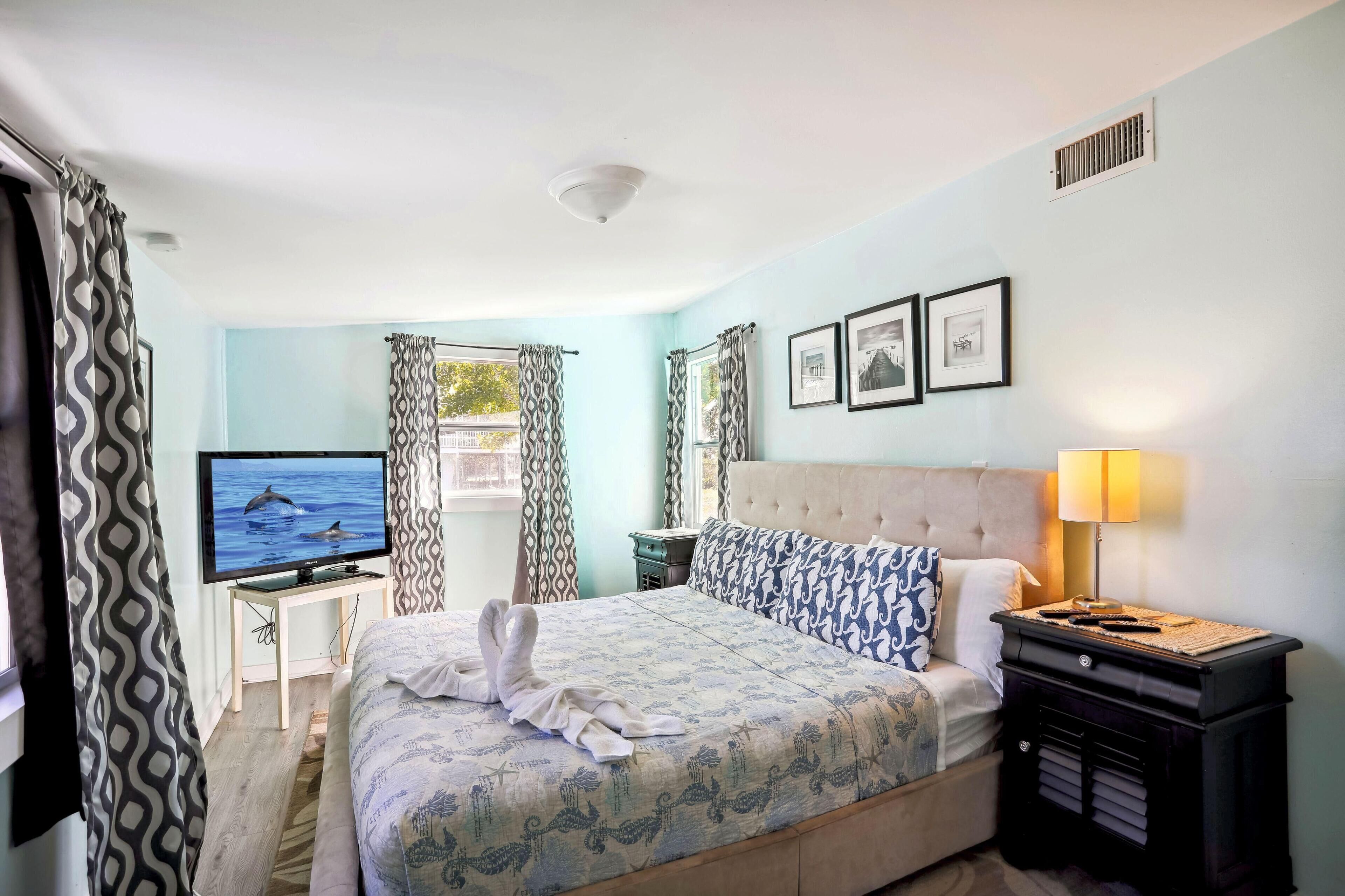 Habitación de hotel con cama, televisor, mesita de noche con lámpara y ventana con cortinas.