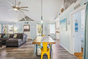 A spacious living room with a wooden dining table, a ceiling fan, and a large window. - Beach Life 3 (Treasure Island)