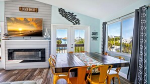 A dining area with a wooden table and yellow chairs, a fireplace, a TV displaying a sunset, and a view of the beach through the window. - Beach Life 5 (Treasure Island)