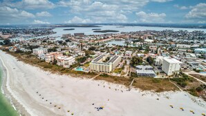 Vue aérienne d’une ville côtière avec une vaste plage, plusieurs bâtiments et un port de plaisance.
