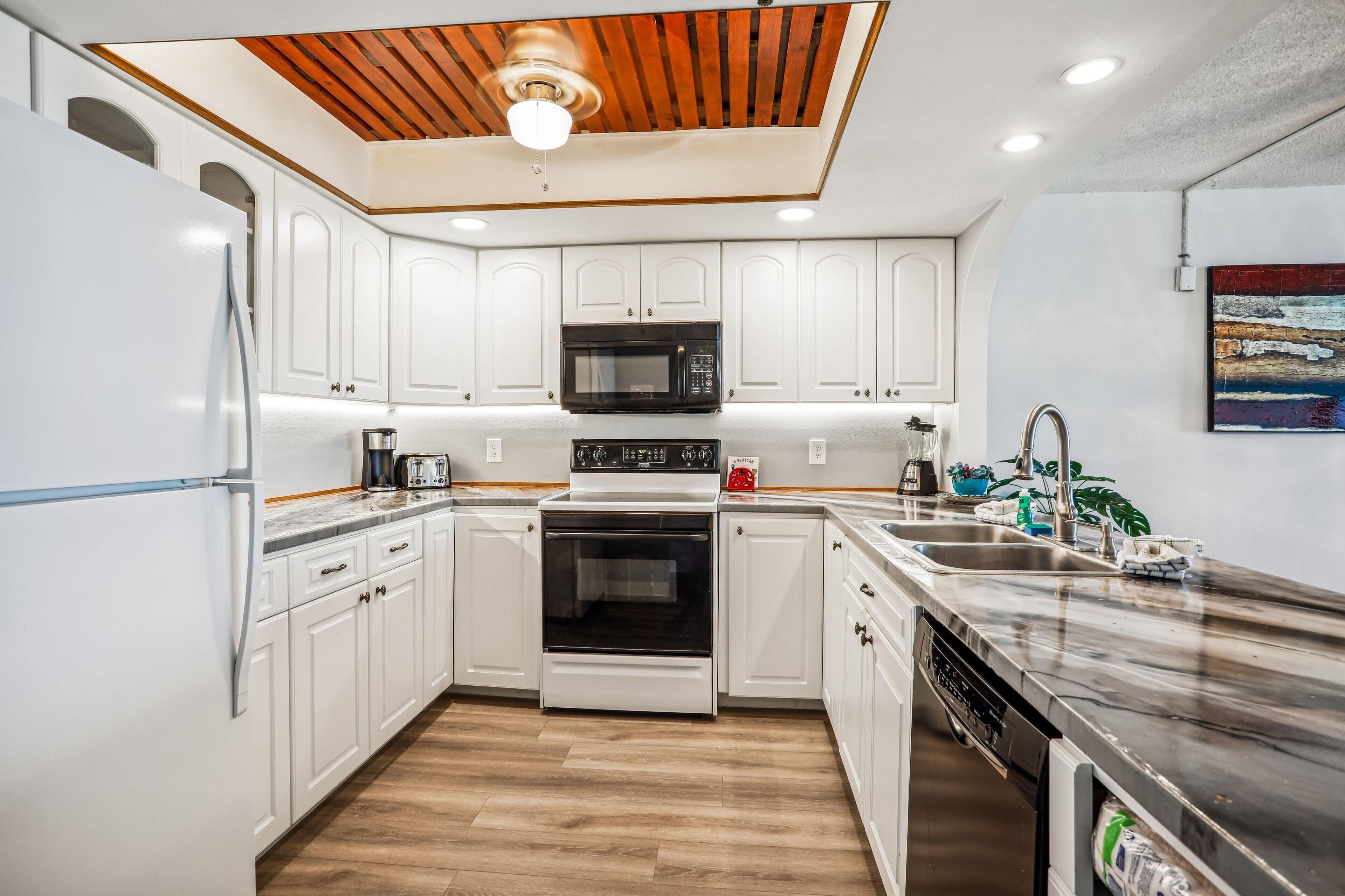 A modern kitchen with white cabinets, a black microwave, a stainless steel sink, and a wooden ceiling.