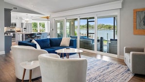 A spacious living area with a large window overlooking a body of water, a kitchen in the background, and a balcony with outdoor seating.