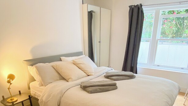 Una cama bien hecha con almohadas y una toalla doblada, una mesita de noche con lámpara, y una ventana con cortinas.