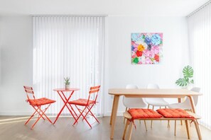 Ein heller Raum mit einem Esstisch, roten Stühlen, einer Bank und einem farbenfrohen Blumenbild an der Wand.