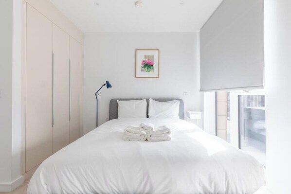 A modern bedroom with a large bed, a bedside lamp, and a window with blinds.