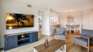 Un salon moderne avec un téléviseur à écran plat, une table basse et des sièges. On aperçoit à l’arrière-plan un coin repas avec une table et des chaises.