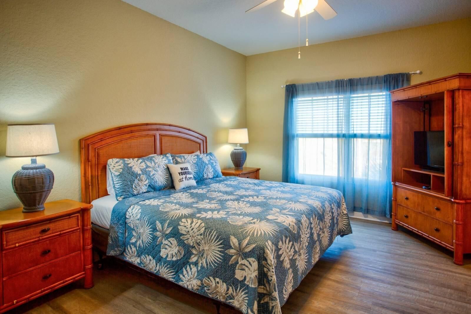 Un dormitorio con una cama, dos mesitas de noche, un armario de madera, un ventilador de techo y una ventana con cortinas.