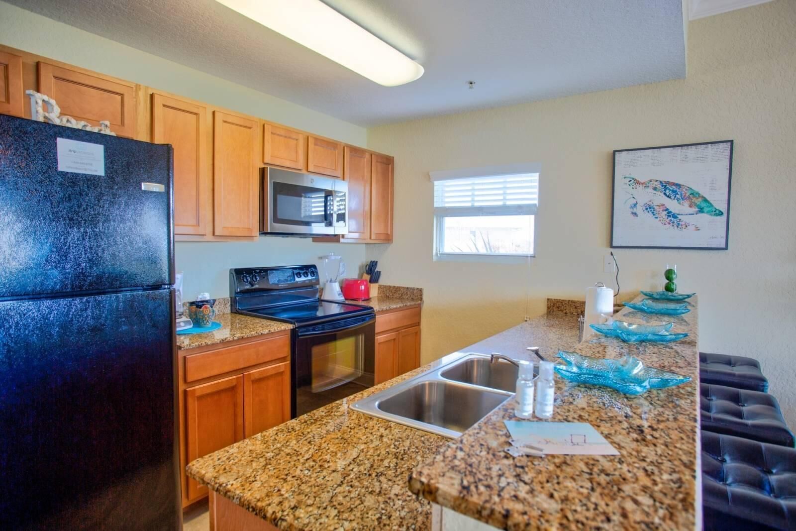 A kitchen with granite countertops, wooden cabinets, a black refrigerator, a microwave, a stove, and a sink.