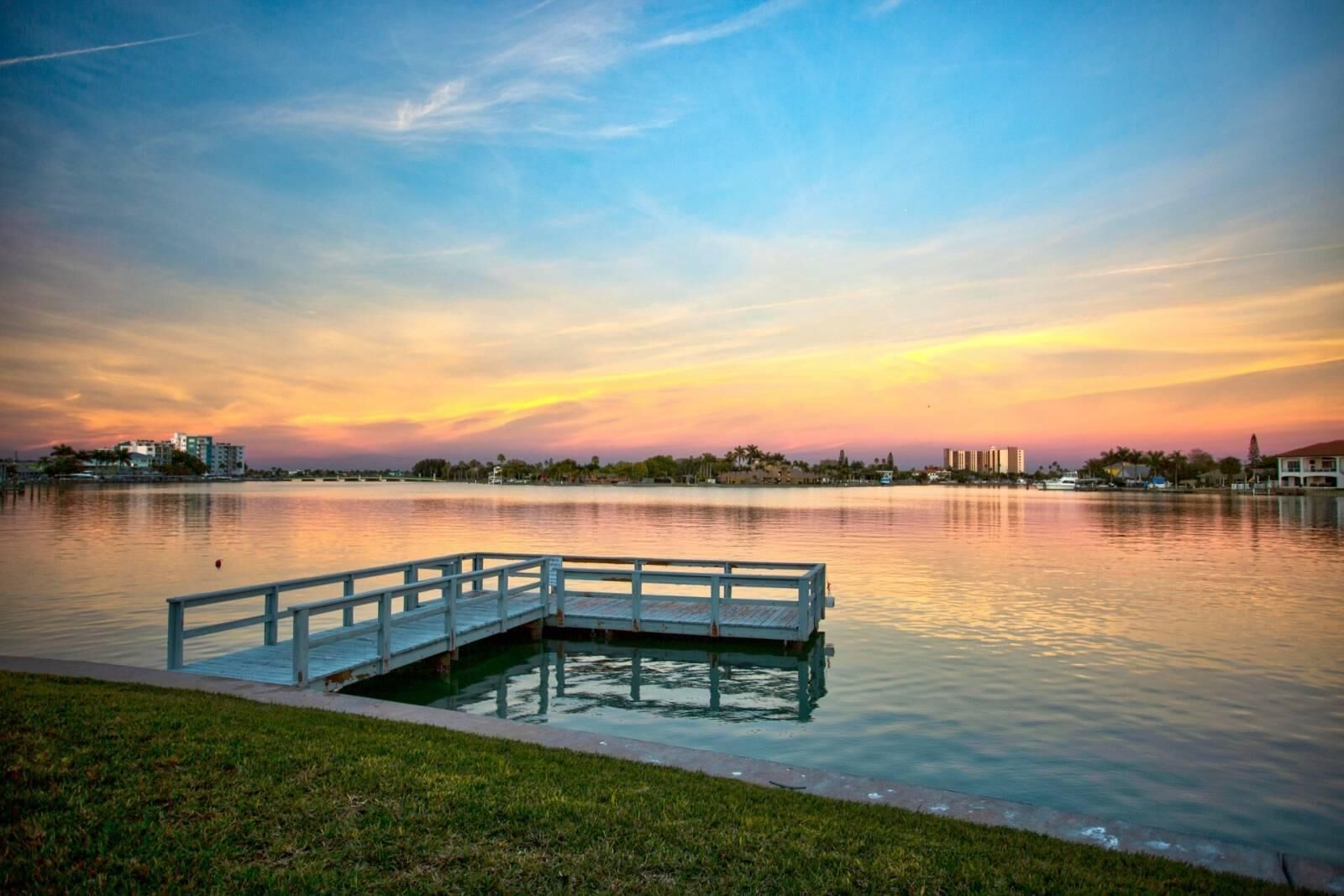 Un parque frente al agua con un muelle de madera que se adentra en aguas tranquilas, rodeado de zonas verdes y edificios al fondo.