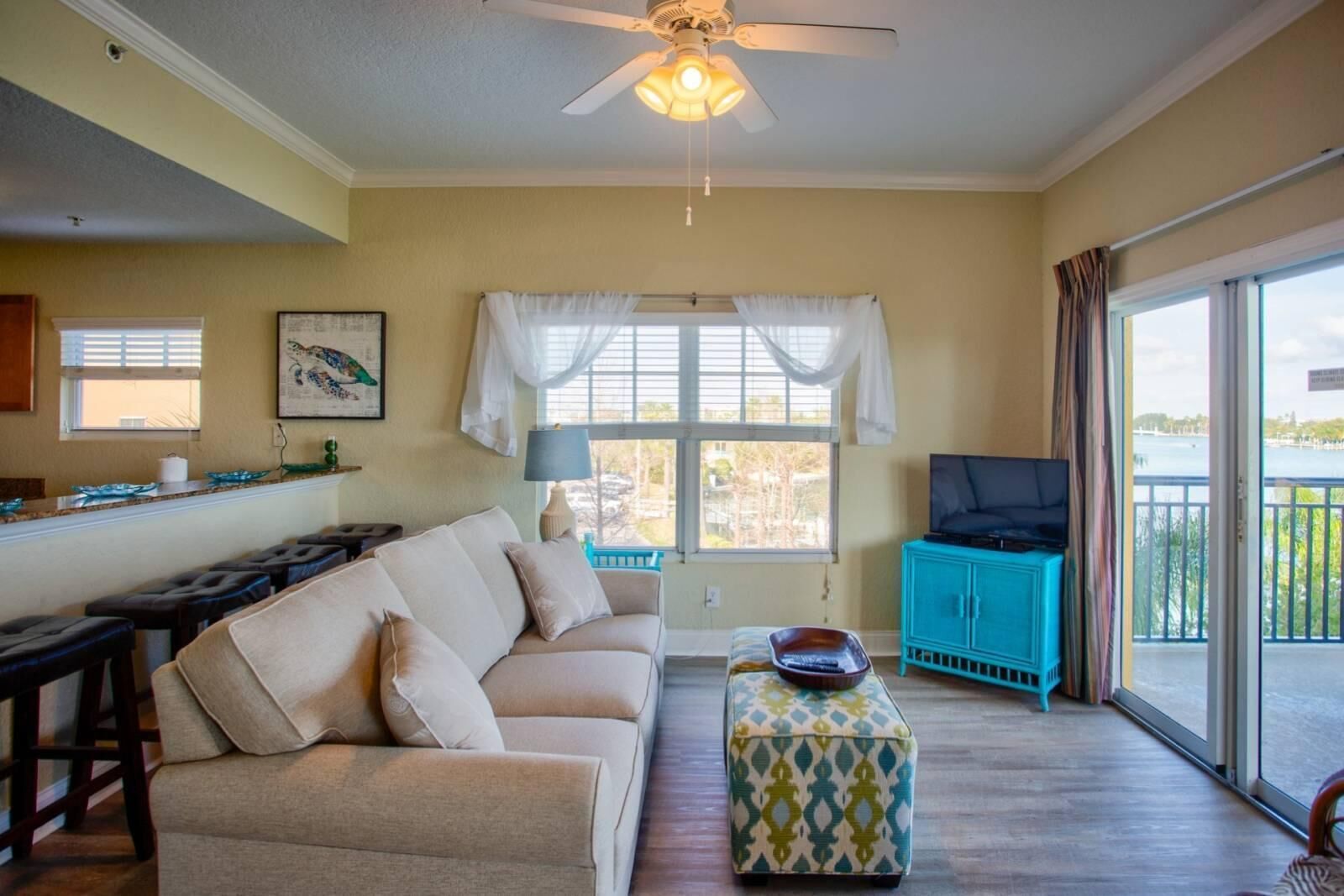 A living room with a sofa, ottoman, and a television on a stand. There is a balcony with a view of the outdoors.