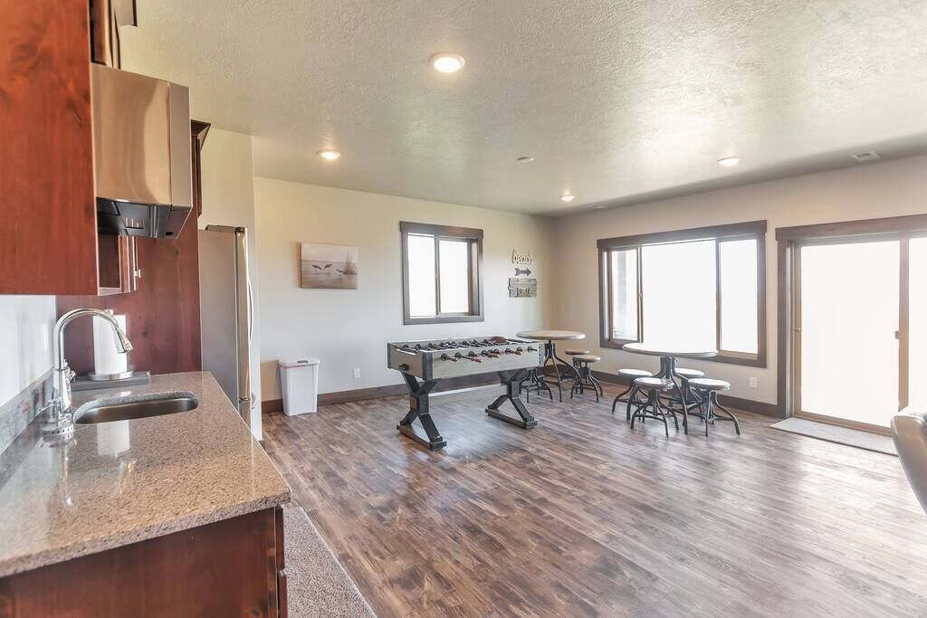 A spacious kitchen with a foosball table, a dining area with a round table and chairs, and large windows providing natural light.