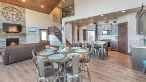 A spacious living area with a fireplace, a dining table with chairs, a kitchen with modern appliances, and a staircase leading to an upper level.