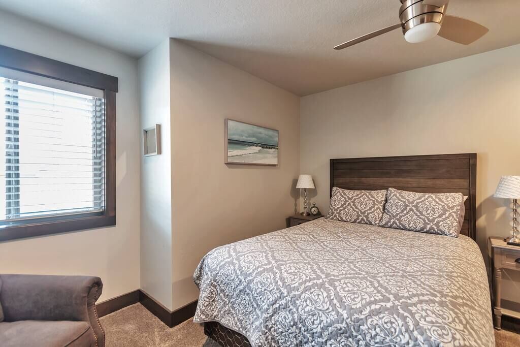 Un dormitorio con cama, mesita de noche, lámpara, silla, ventilador de techo y ventana con persianas.