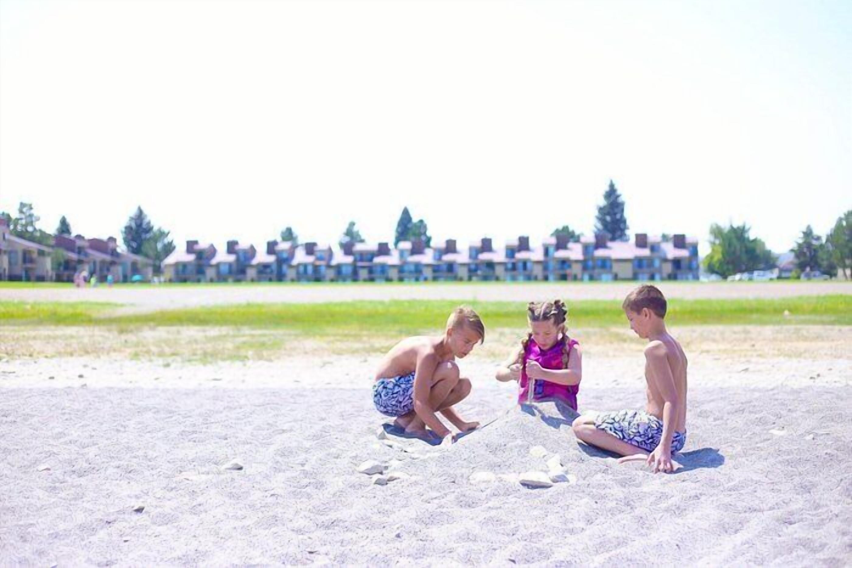 Tres niños jugando en una playa arenosa con una hilera de casas al fondo.