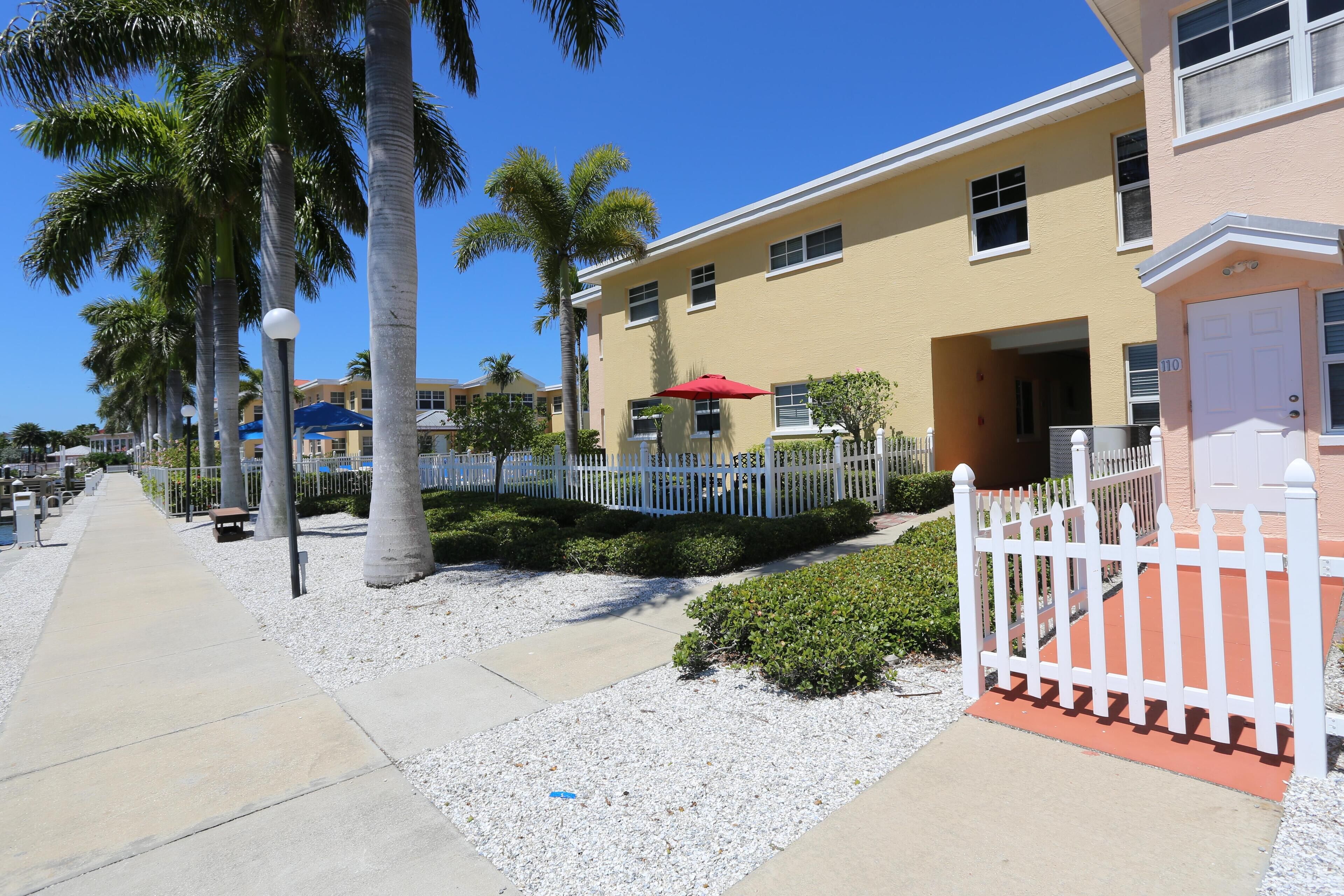A residential building with a white picket fence, palm trees, and a red umbrella.