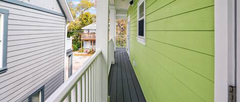 A narrow wooden deck with white railings, connecting two houses with green siding and white trim.