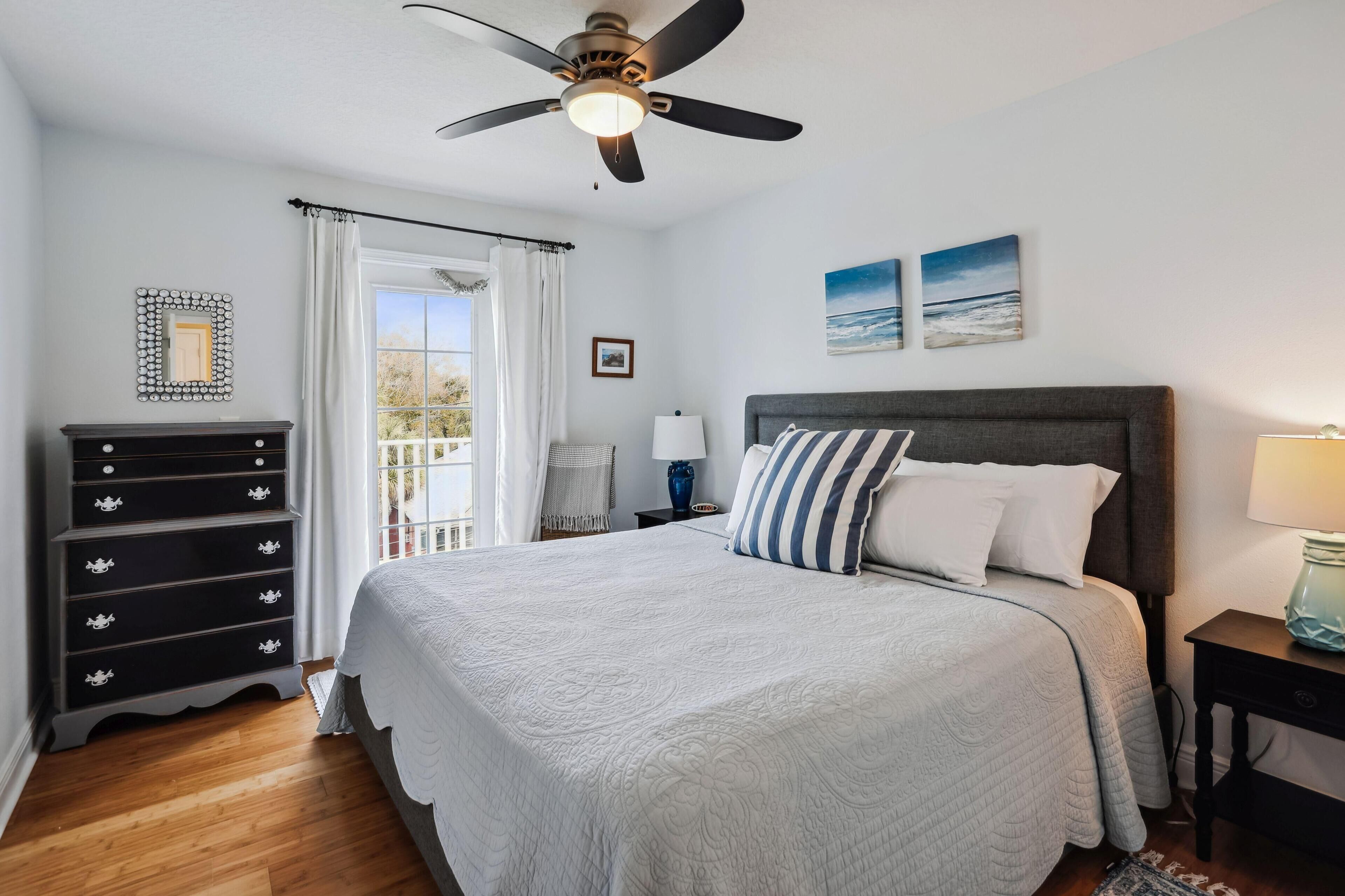 Un dormitorio con cama, cómoda, ventilador de techo, ventana con cortinas y dos cuadros colgados en la pared.