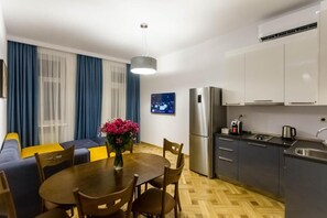 A compact kitchen with modern appliances, a dining area with a wooden table and chairs, and a sofa with blue and yellow cushions.
