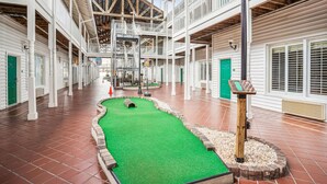 Um espaço amplo e coberto com campo de mini golfe em grama sintética verde, guarda-corpos de madeira branca e teto alto com vigas aparentes.