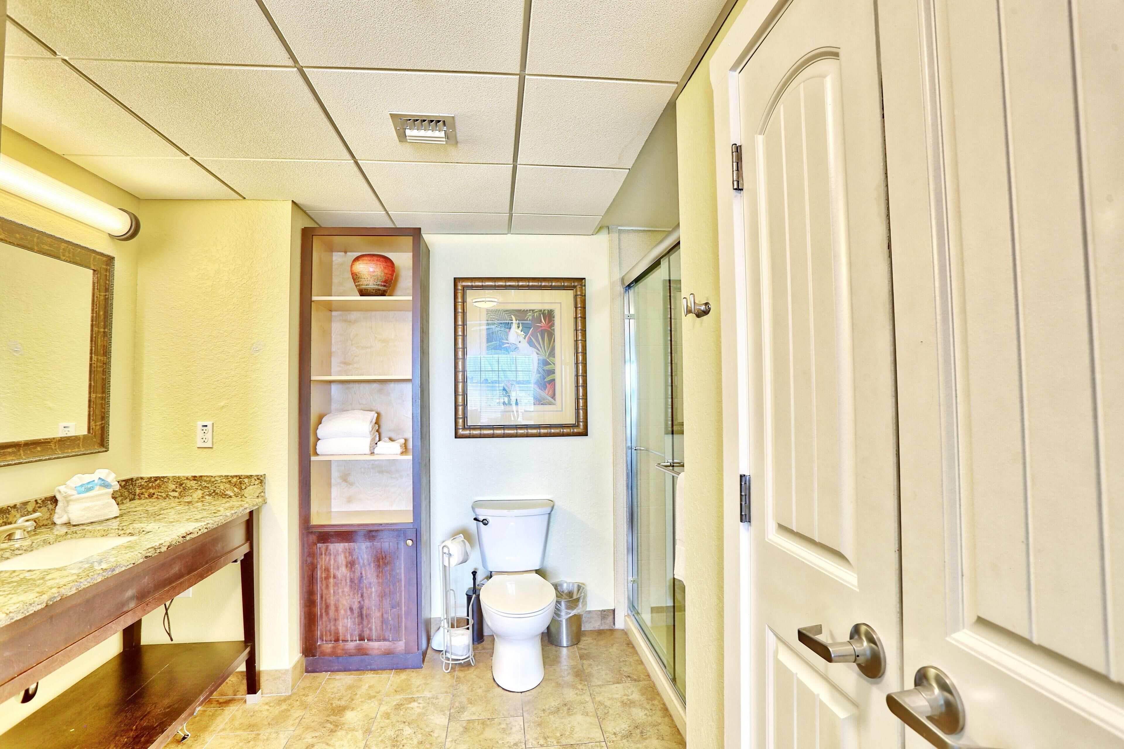 A bathroom with a marble countertop, a wooden vanity, a toilet, a framed picture on the wall, and a door with a silver handle.