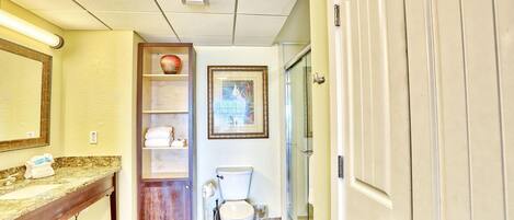 A bathroom with a marble countertop, a wooden vanity, a toilet, a framed picture on the wall, and a door with a silver handle.