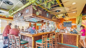 Uma área de bar com banquetas de madeira, um balcão com caixa registradora e diversos utensílios de bar.