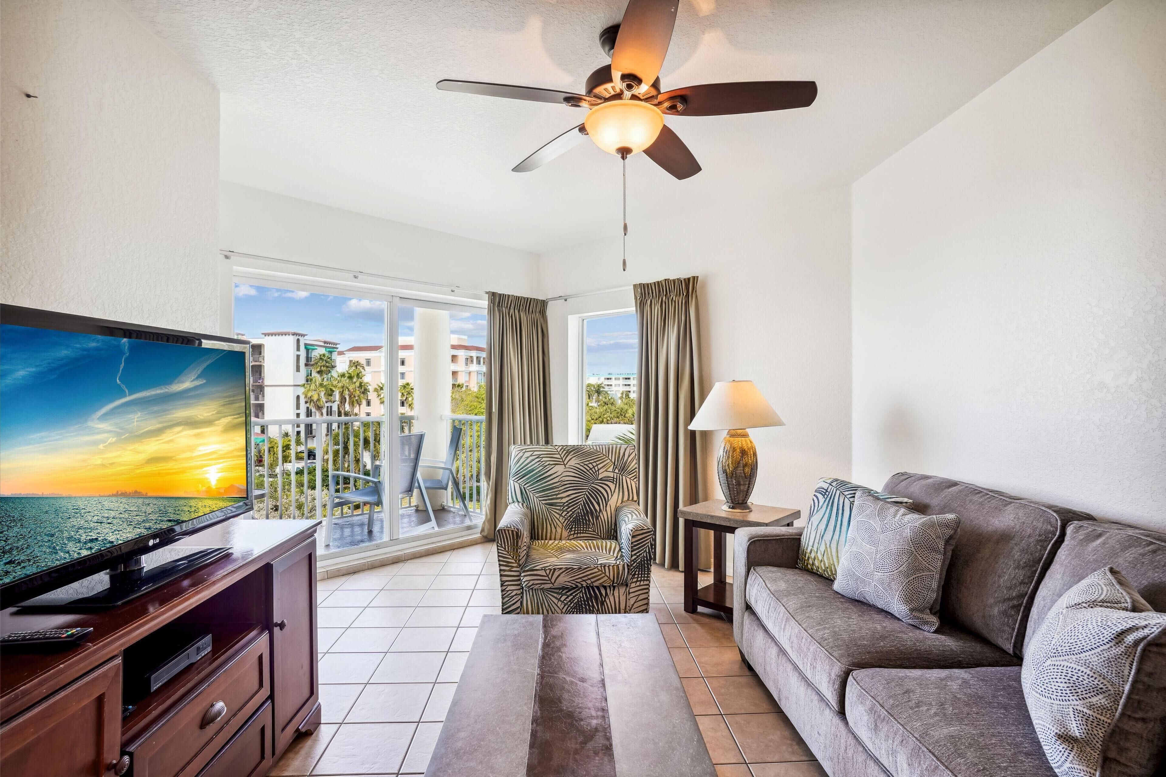 A modern living room with a flat-screen TV, a ceiling fan, a sofa, a patterned armchair, and a view of the ocean through large windows.