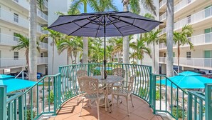 Un balcon avec une table et des chaises, entouré d’une balustrade, avec des palmiers et un immeuble en arrière-plan.