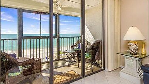 A balcony with a view of the beach, a glass door, a chair, and a table with a lamp.