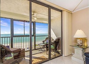 A balcony with a view of the beach, a glass door, a chair, and a table with a lamp. - Surfside South #312 (Madeira Beach)