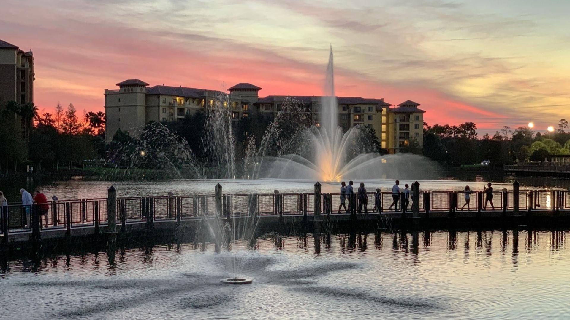 Une vue au coucher du soleil d’une fontaine avec des jets d ’eau, entourée d’une clôture et avec des bâtiments en arrière-plan.