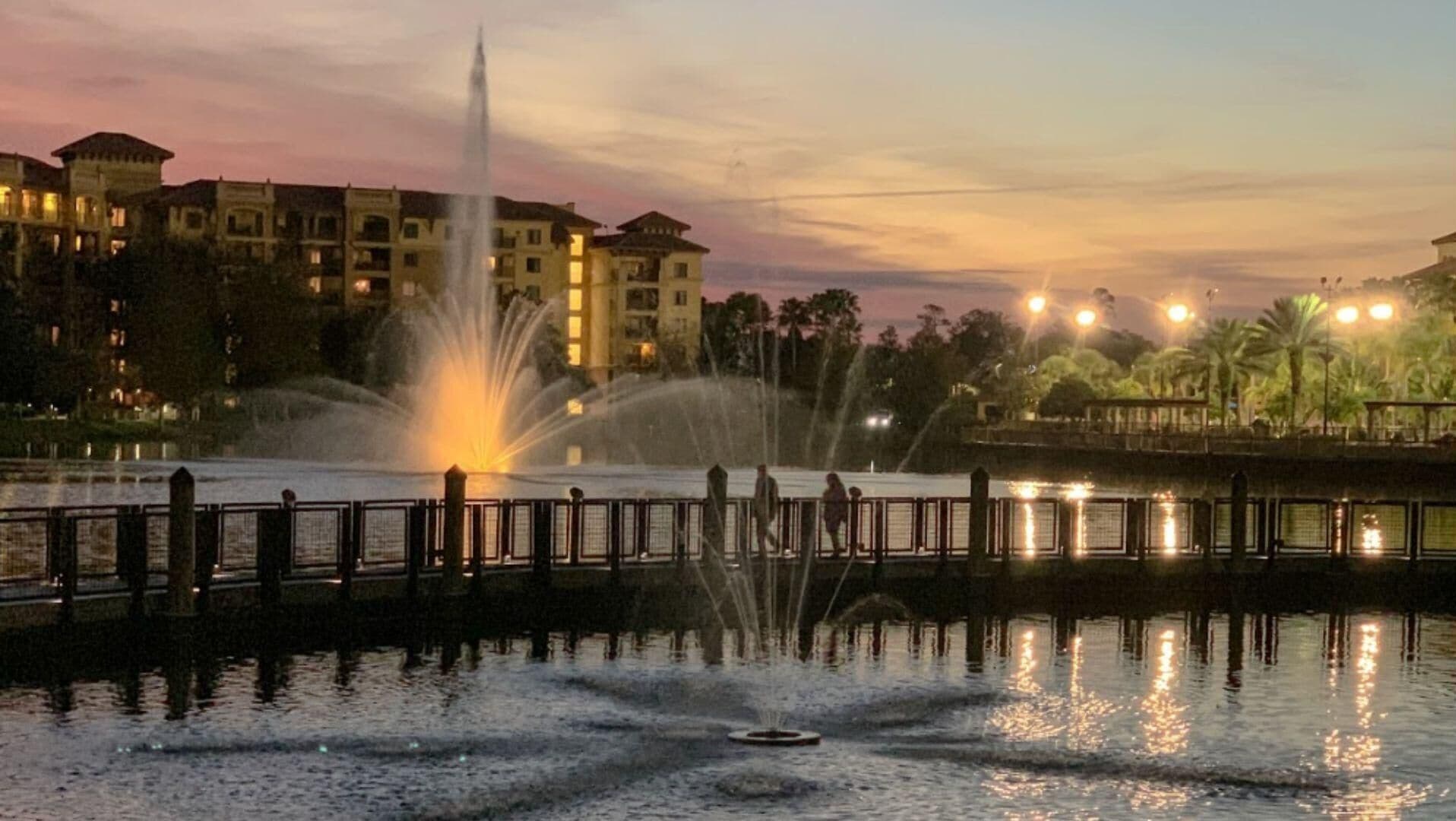 A fountain in a park with a reflection in the water, surrounded by a fence and buildings in the background.