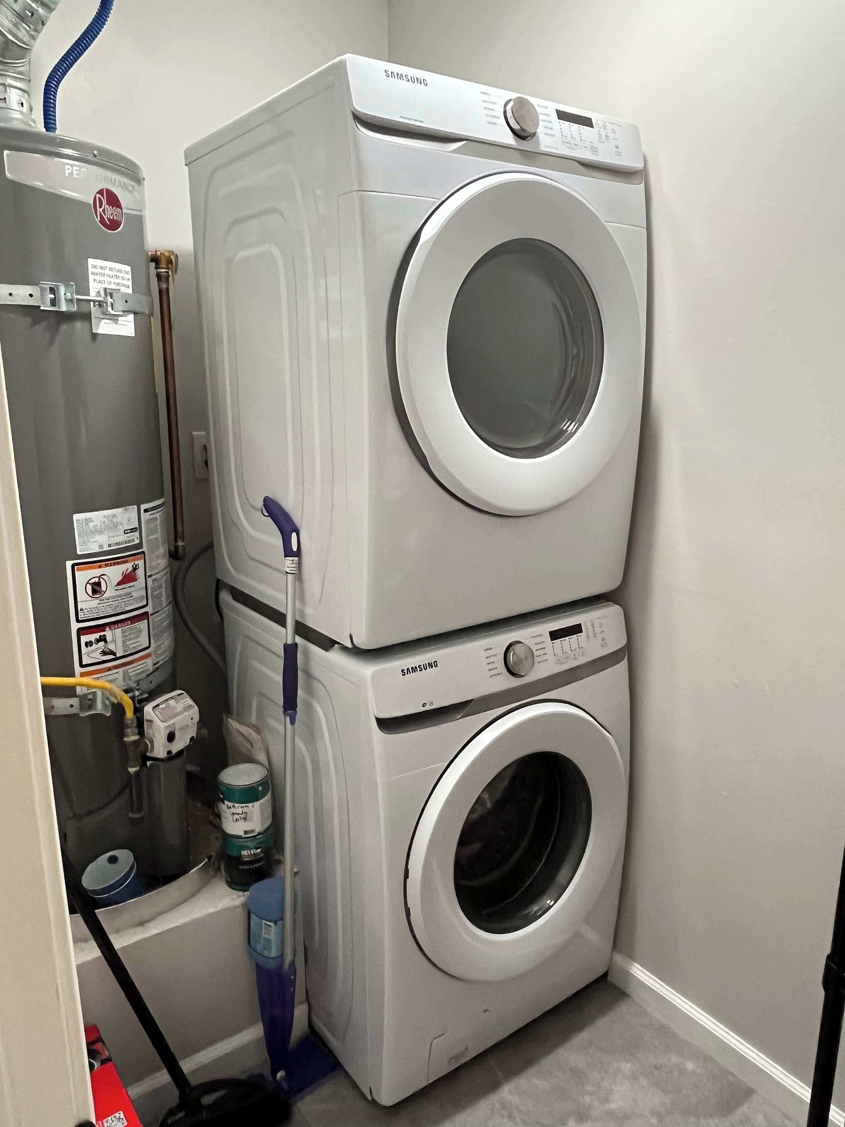 A compact laundry room with a stacked washer and dryer, a water heater, and a hose.