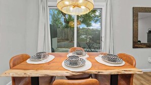 Un coin repas avec une table en bois dressée pour quatre personnes, un grand luminaire tressé suspendu au-dessus et une vue sur un jardin, visible à travers une porte-fenêtre coulissante.