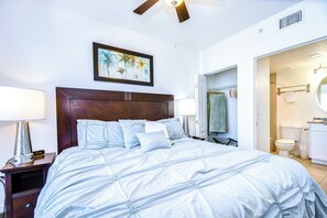Un dormitorio con una cama grande, una mesita de noche con lámpara, un ventilador de techo y un cuadro en la pared.