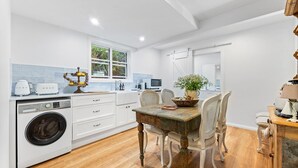 A modern kitchen with white cabinetry, a built-in washing machine, and a dining area with a rustic table and chairs. - Pipis Pretty Point - Couples Retreat (Malua Bay)