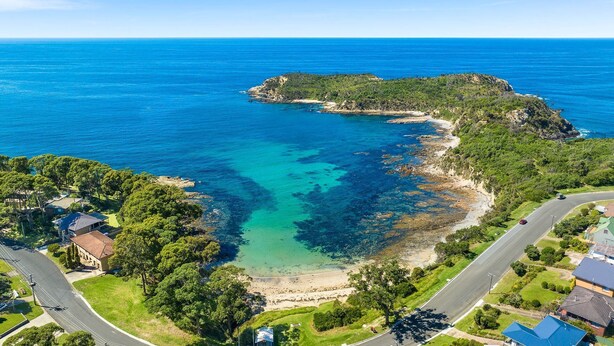 An aerial view of a coastal area with a clear blue sea, a small island with dense vegetation, and residential buildings along the coastline. - Pipis Pretty Point - Couples Retreat (Malua Bay)
