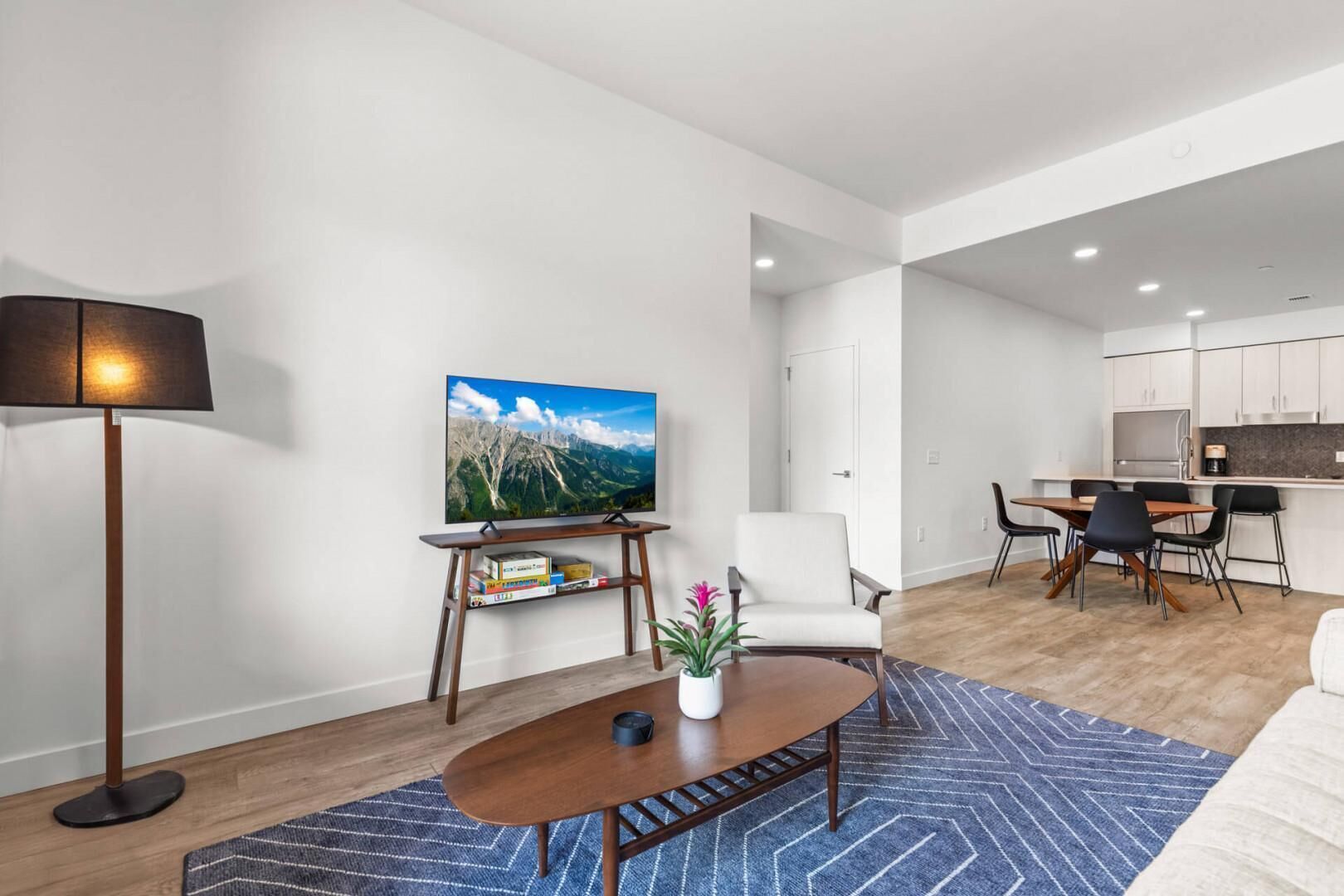 A modern living room with a flat-screen TV, a coffee table, and a dining area with chairs and a kitchen in the background.