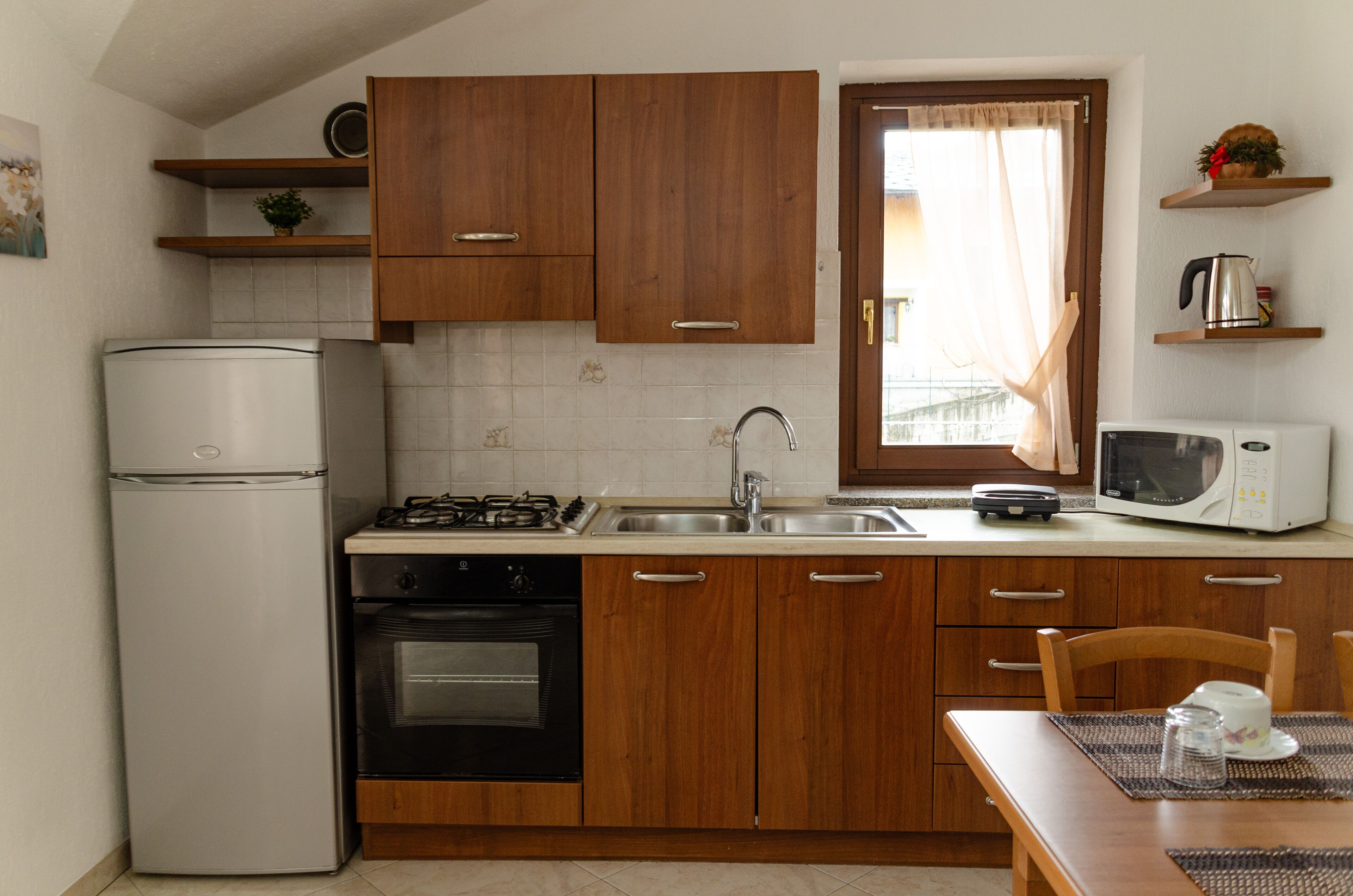 A compact kitchen with wooden cabinets, a refrigerator, stove, oven, microwave, and a window with curtains.