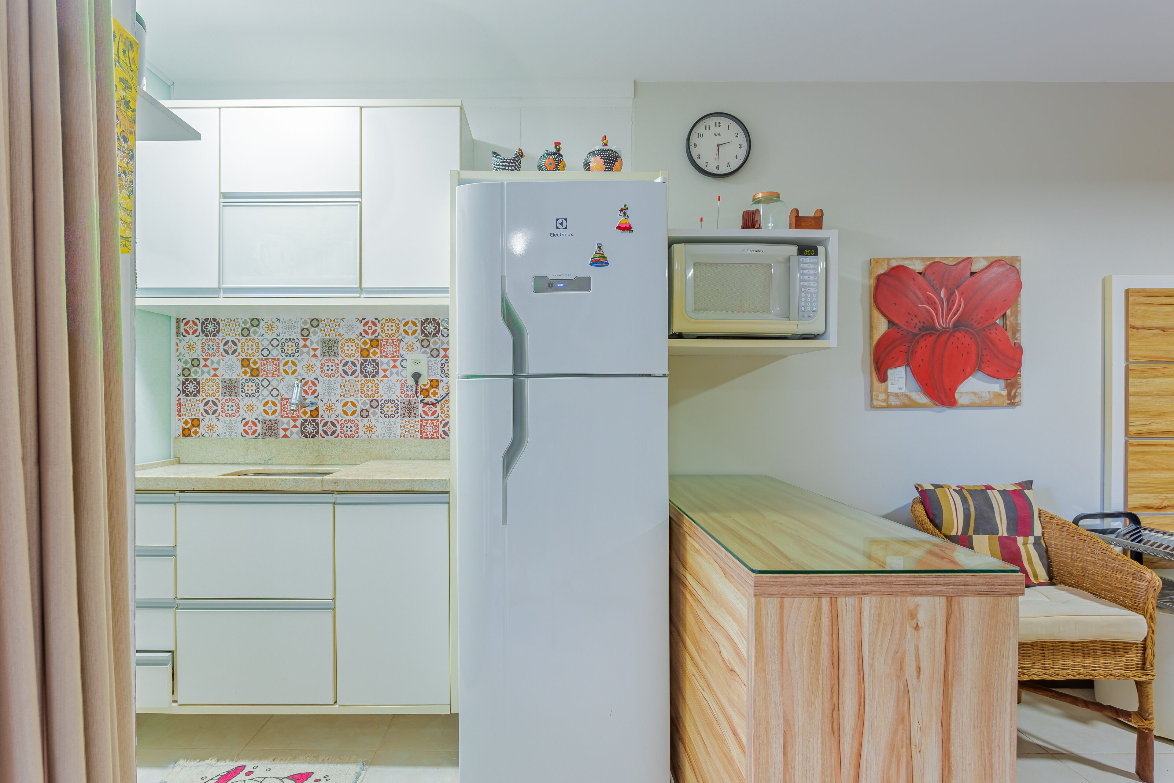 A compact kitchen with white cabinets, a refrigerator, a microwave, and a dining area with a glass table and wicker chair.